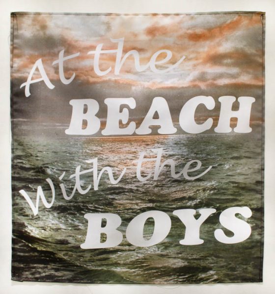 A piece of fabric with a print of ocean and a cloudy sky at golden hour with the words: At the beach with the boys