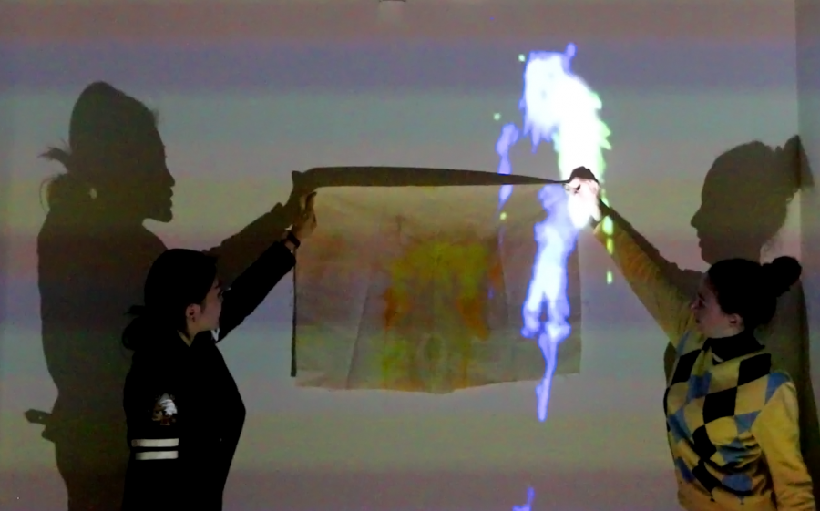 Two girls are standing next to a wall holding a piece of fabric with an abstract painting, and an image is projected on the wall casting shadows of them