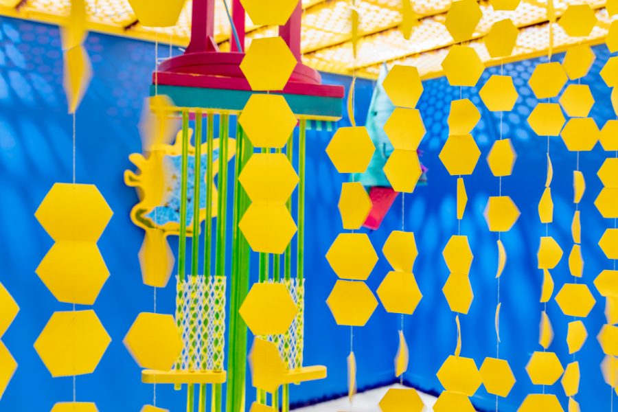 An installation view of artwork by Juliette Sardou. Multicolored sculptures using multiple materials placed in front of a blue wall. One sculpture is hanging from a ceiling in the center of the space. There is yellow material with hexagonal cutouts draped across the ceiling. Yellow hexagonal shapes attached to thread in the foreground.