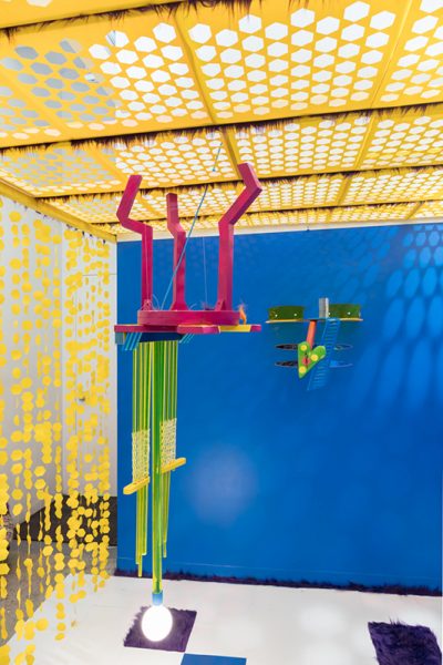 An installation view of artwork by Juliette Sardou. Multicolored sculptures using multiple materials placed in front of a blue wall. One sculpture is hanging from a ceiling in the center of the space. There is yellow material with hexagonal cutouts draped across the ceiling.