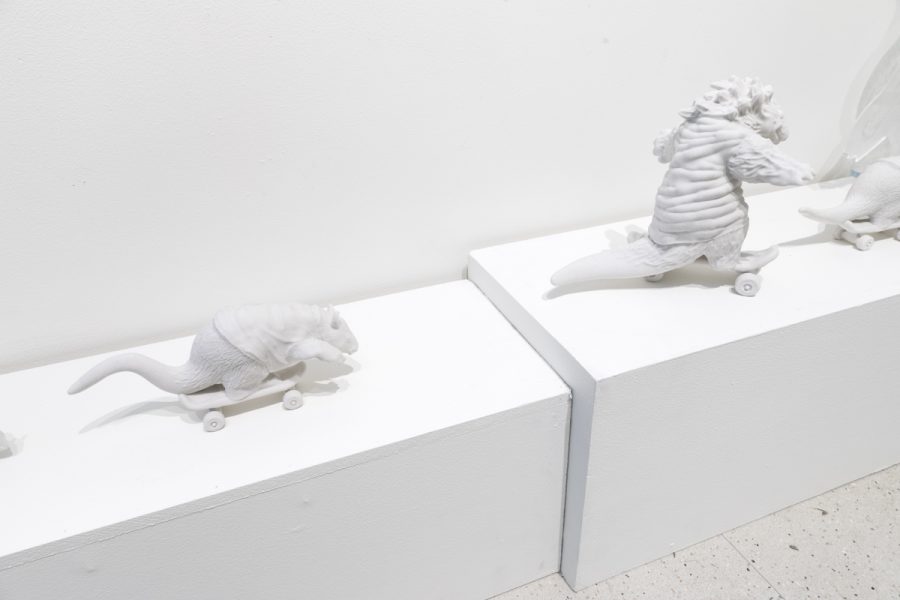 Sculpture by Julio Cesar Candelario. Multiple white sculptures of animals wearing clothes riding a skateboards.