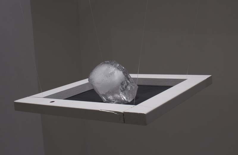 A white wood frame with a black bottom hung on the ceiling with invisible wire holds an organic rounded-shaped silver sculpture.