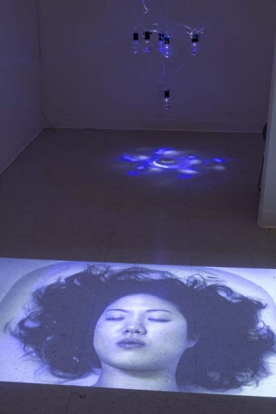 Installation view of aquarium lightbulbs with blue light cast on the floor and a large image projection projected on the floor of a woman's portrait with her hair spread and eyes closed.