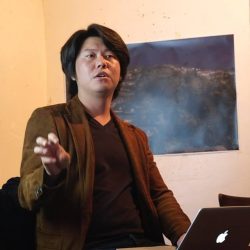 Juno Seo looking in the distance with his arm gesturing and a macbook on his lap, there is a poster of a landscape behind him on the wall