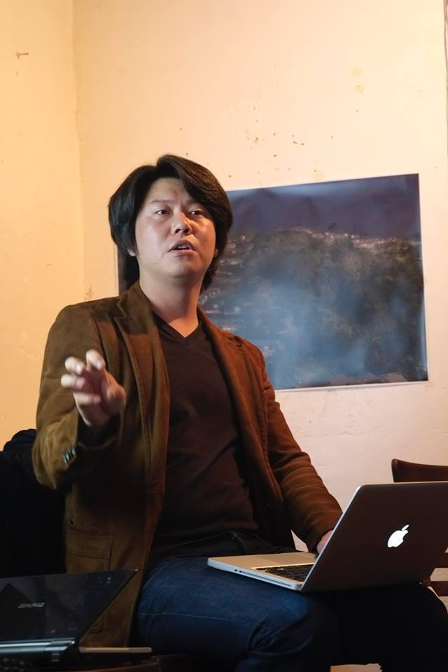 Juno Seo looking in the distance with his arm gesturing and a macbook on his lap, there is a poster of a landscape behind him on the wall