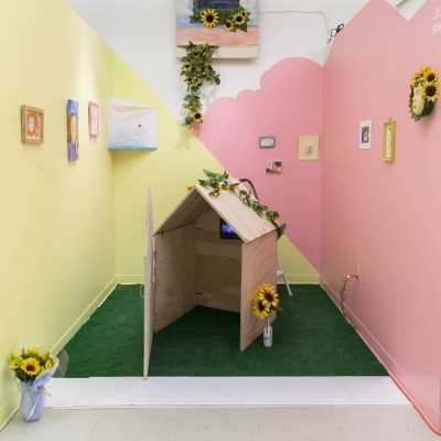 Installation view of a wooden house on a green carpet, yellow and pink walls, paintings hanging on the walls, and many sunflowers on the house, on the wall, and in a vase.