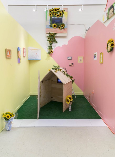 Installation view of a wooden house on a green carpet, yellow and pink walls, paintings hanging on the walls, and many sunflowers on the house, on the wall, and in a vase.