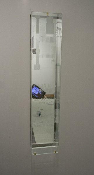 A mirror is installed on the wall, in the shape of a tall rectangular with vinyl shapes installed on it a few words, all in white color, and an image with a laptop and a table are reflected in the mirror.
