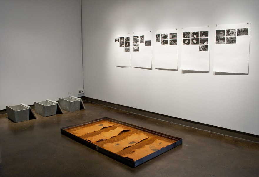 Installation view of a metal tray with paper stripes on a rusted surface, three concrete trays with lids open, and five whiteboards with arrays of black and white photographs