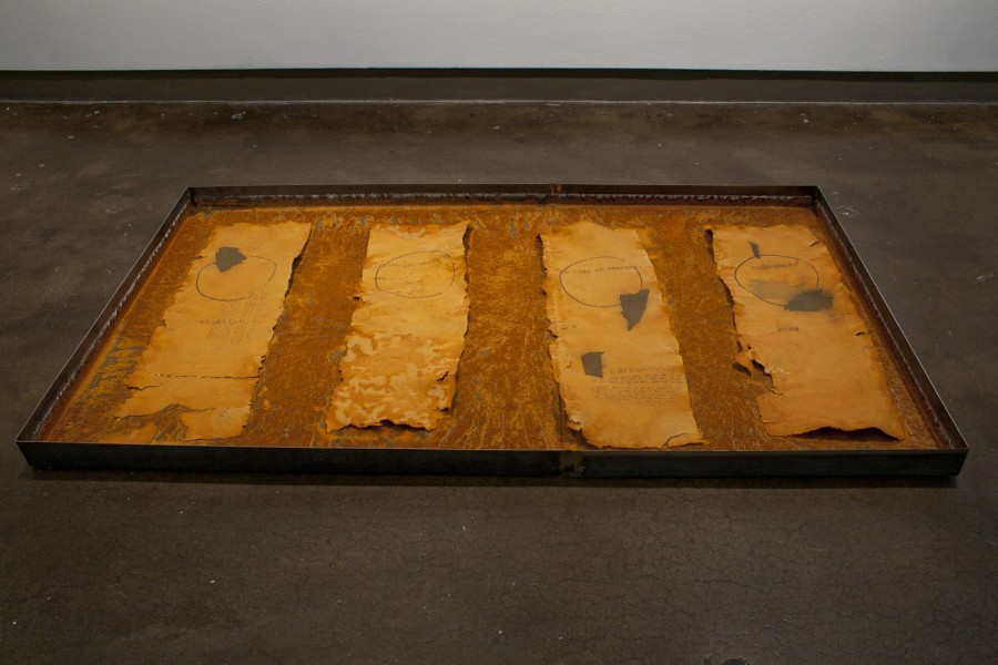 Four orange paper fragments with text, circles and flat shapes displayed on an rusted large metal tray installed on the floor.