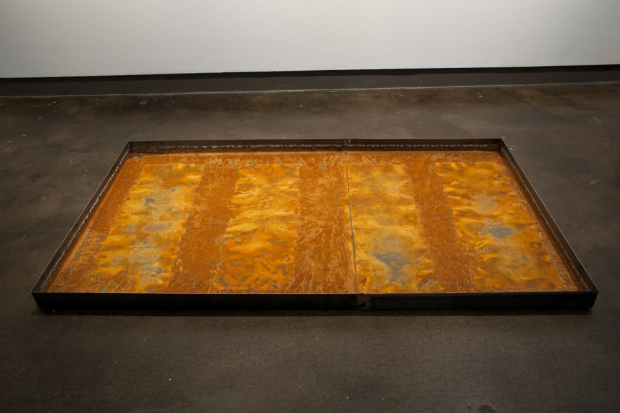 Four orange paper fragments with text, circles and flat shapes displayed on an rusted large metal tray installed on the floor.