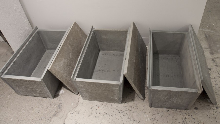 Three concrete-made crates with lids open on their right side, placed on the floor near a white wall.
