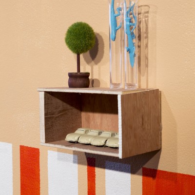 Close view of a wooden box installed on a wall. Inside it is four beige cars, and on top of the box is a small green tree and two blue reptile shapes. Under the box are many stripes in red and white in different dimensions.