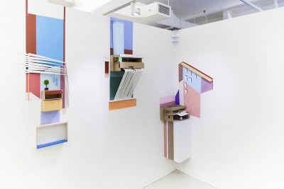 Installation view of mixed media made of cardboard and colorful paper. On the left is a small green tree sitting on a small box with a blue, red, and pink background, in the middle is a cardboard structure on a blue, red, and green background, and on the right is another cardboard structure with a pink, blue and violet background and the shapes of windows of a building made in the scene.