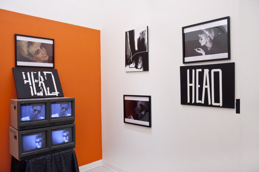 Photographs in black frames mounted on an orange and white wall and an array of four TVs with portrait images displayed.