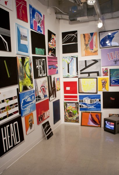 Exhibition view of many paintings illustrating portraits, abstract shapes, text messages, mounted in the corner, and a small TV on the floor with an image displayed.