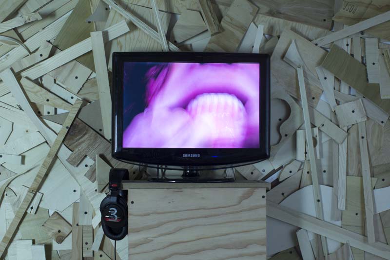 A wall made from different sized wood pieces and installed with nails, a wood stand, and a TV with a distorted image of a mouth with teeth visible in a pink light