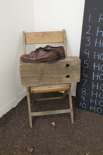A wood chair in a corner with a big wood piece which has a pair of man's brown shoes and a part of a blackboard on the right side with a ten-item list