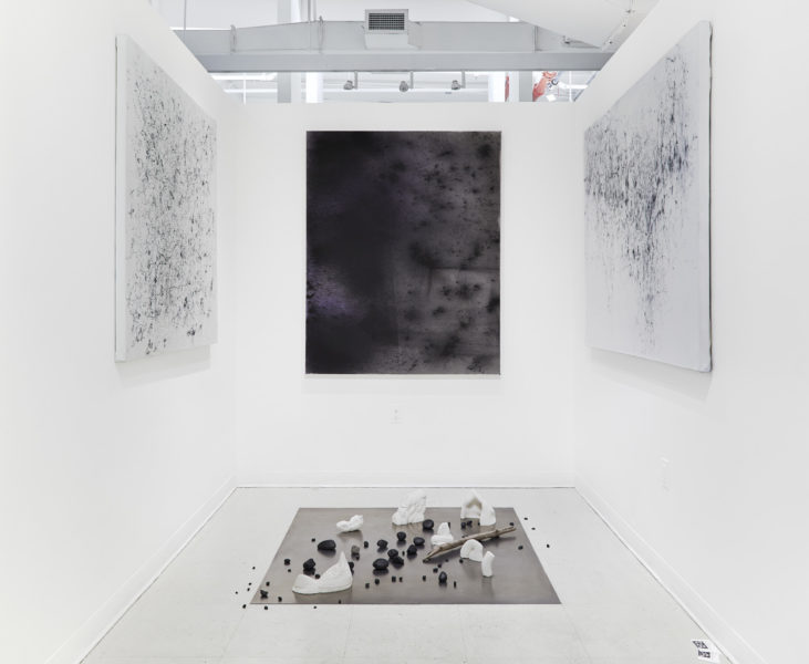 An installation of black and white drawing on canvas with several objects arranged on the floor, in a white gallery space.