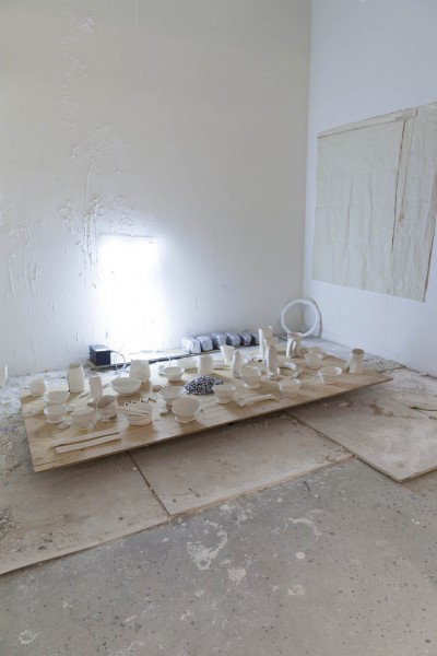 Installation view of ceramic bowls, cans, and glasses placed on a wooden floor and illuminated from the wall in the background.