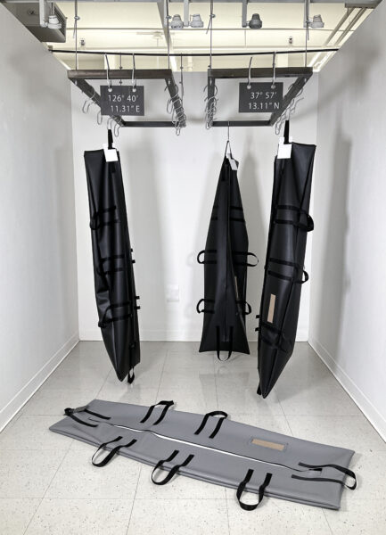 Three black body bags hanging upright with hooks attached to two separated metal racks and one grey body bag placed on the floor