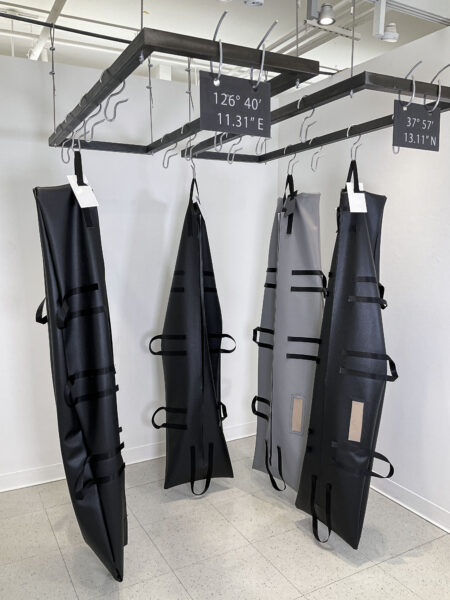 Three black body bags and a grey body bag hanging upright with hooks attached to two separated metal racks