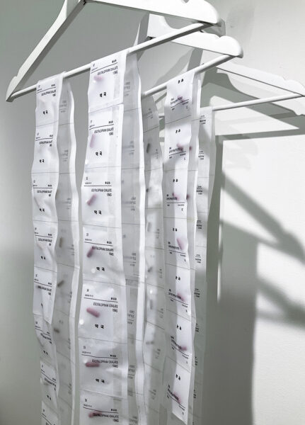 Contiguous white strips of packets containing pills and labelled with black text hang from three wooden clothes hangers painted white.