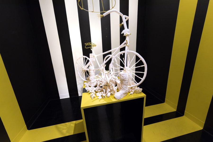 Installation view of artwork by Kirin Pino. Various unglazed ceramic figures and objects placed on a yellow pedestal. In the center is a fabricated white bicycle. The walls have yellow, white and black stripes ion them.