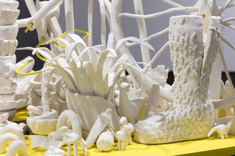 Detail of multiple sculptures by Kirin Pino. Various unglazed ceramic figures and objects placed on a yellow pedestal.