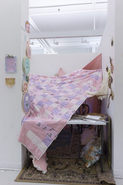 Installation view of pink blanket partially covering a desk and a chair in a corner and many other colorful embroilments hanging on the wall