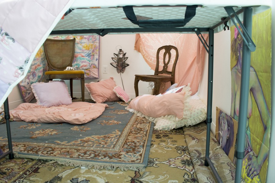 Installation view of two chairs and many orange and green pillows sitting on the carpets under an artisanal tent and representation of nude woman on a piece of fabric on the side.