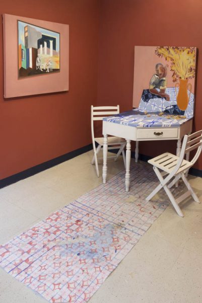 Installation view of a white hexagonal table with two white chairs, a painting with a kid sitting on the table next to a vase with flowers, a white carpet with red diamond shapes, on the floor, and two red walls and on one of the wall is hung a painting with an architectural construction