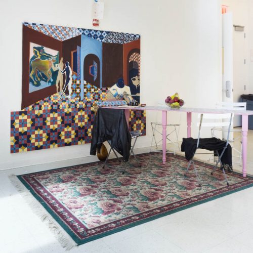 Installation view of a dining table with pink legs, four white chairs and two of them hold a black leather jacket and one a scarf, on the floor is a flower pattern carper with pink flowers and green leaves, there is a large painting on the wall behind the table representing a large room with a person looking at a painting and another one sitting on a couch playing on the guitar. The room in the painting is with red walls and has a pattern with yellow, red, blue, and black squares