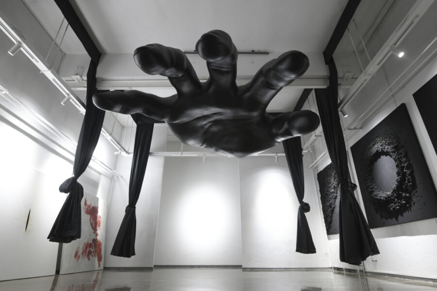 A sculpture of a large black human hand suspended in a white-walled gallery. Black fabric curtains hang from the ceiling, tied of in knots, and black deep relief wall hangings line the wall on the left.
