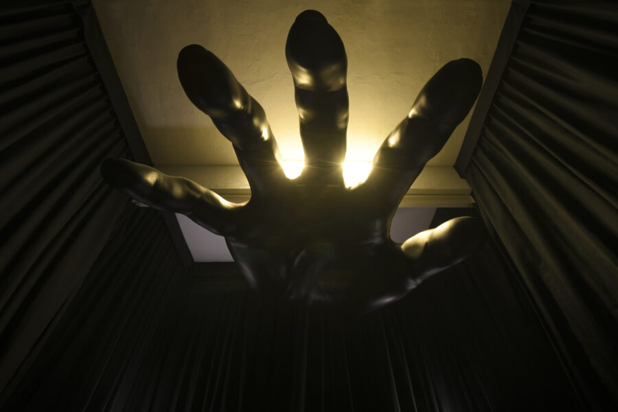A sculpture of a large black hand is suspended from the ceiling, back-lit and shot from below. Black fabric curtains line the walls on all sides.