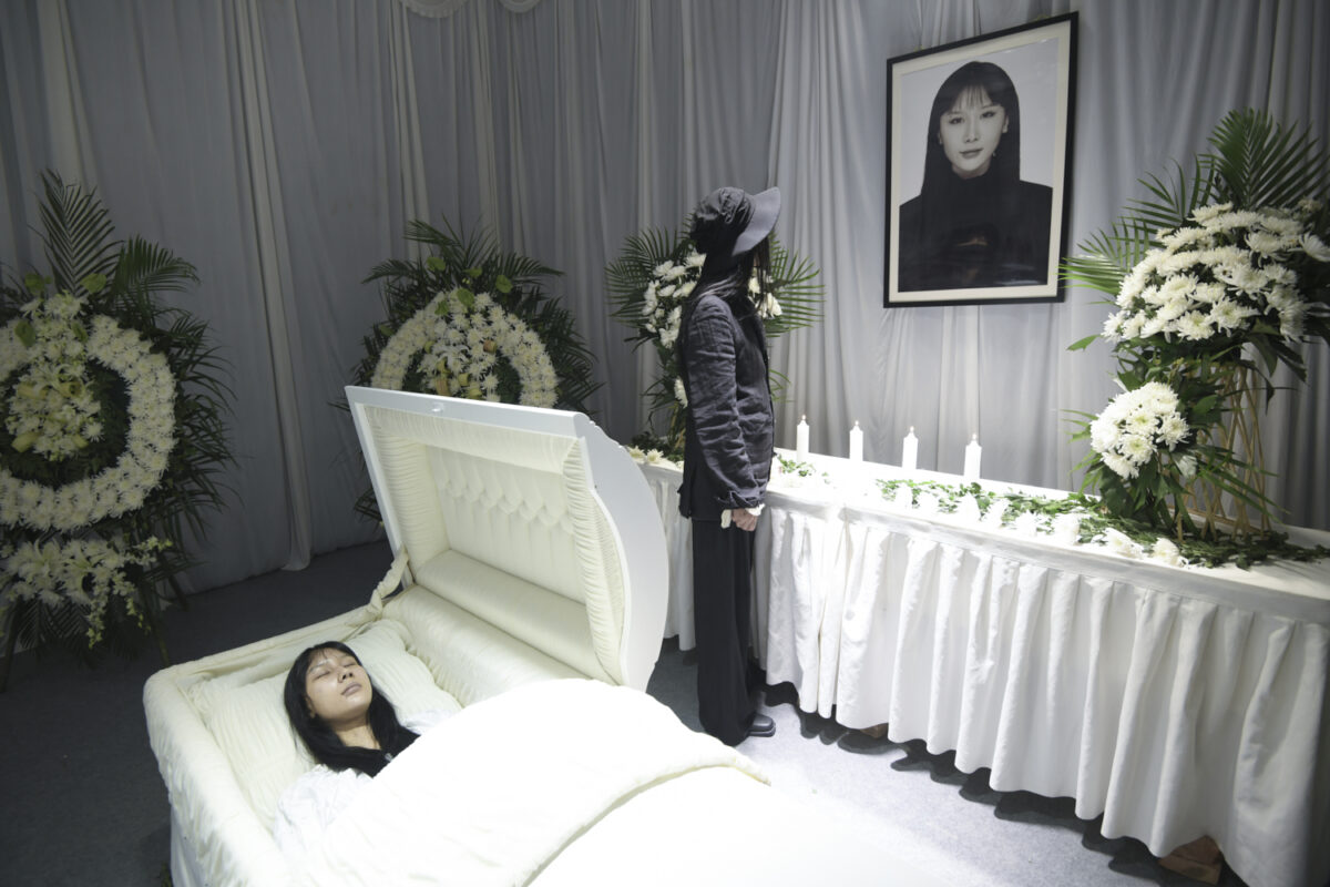 An the artist lies in open white casket in a room draped in white fabric with white flowers and candles. I dark clad mourner whose face is obscured (and may also be the artist) gazes at a photographic portrait of the artist.