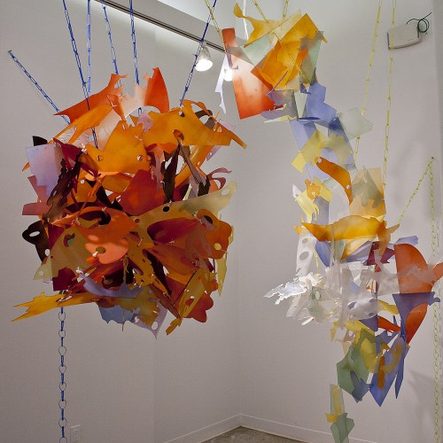 Different angled and rounded shapes made of colored material, including orange, red, blue, white, hung on the ceiling with chains made of blue and yellow fabrics.