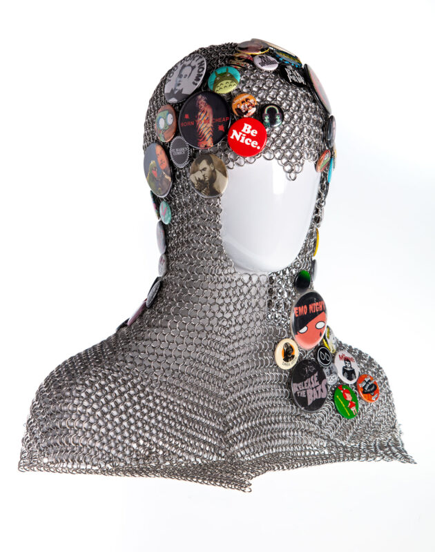 A chainmaille coif displayed on a white plastic mannequin head. The coif is adorned with pinback buttons of various sizes and colors. In this photo the right side of the coif is visible.