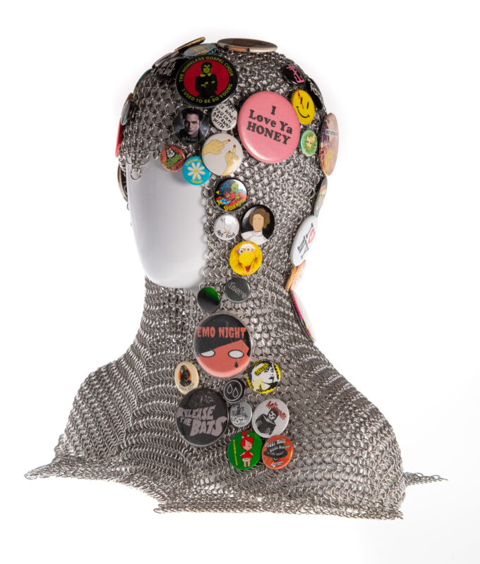 A chainmaille coif displayed on a white plastic mannequin head. The coif is adorned with pinback buttons of various sizes and colors. In this photo the left side of the coif is visible.