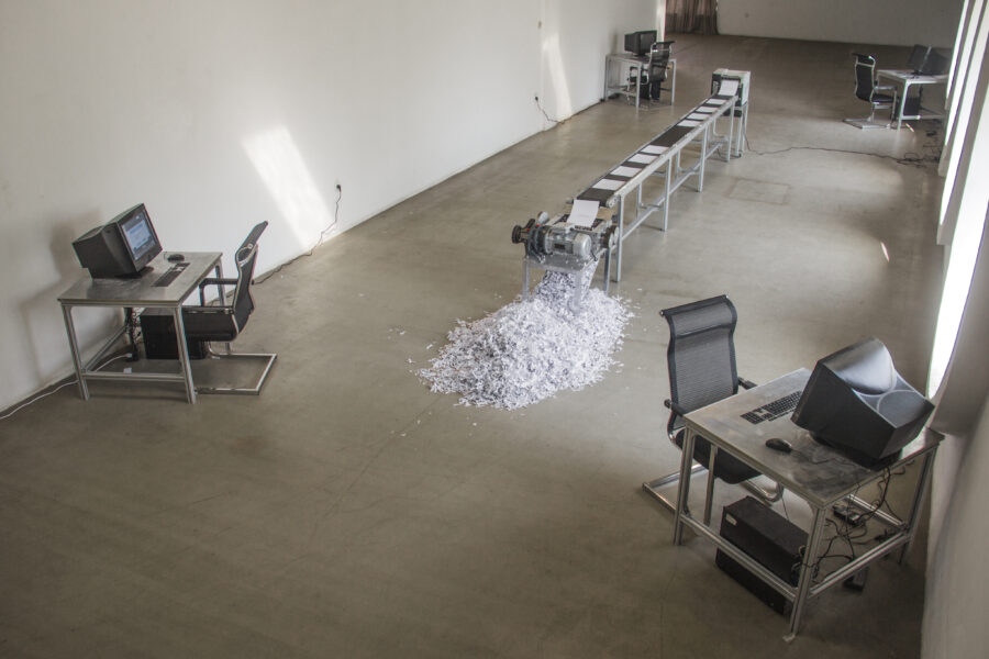 Artwork by Jiaqi Li. The installation shows a room with a conveyor belt and four desks in each corner with a computer and black chair. Each desk faces the wall.