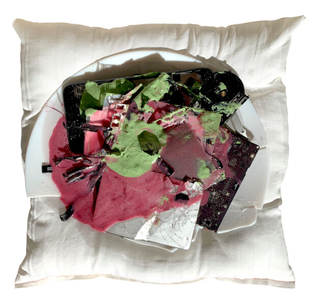 The artwork shows a square white pillow. On top of the pillow is a broken white ceramic plate with unknown objects of brown pink and green.