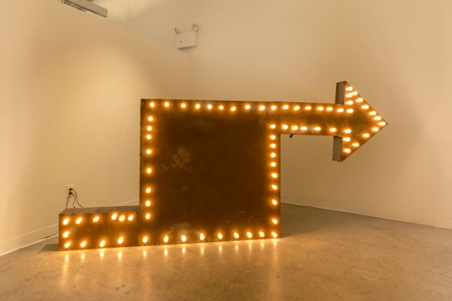 Installation shot of a sculpture that resembles a vintage sign made of metal and yellow Edison light bulbs.