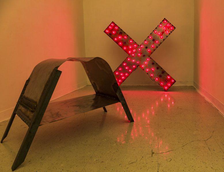 Installation shot of a metal X leaning against the wall, illuminated by red lightbulbs with a metal upside down U form in the foreground.