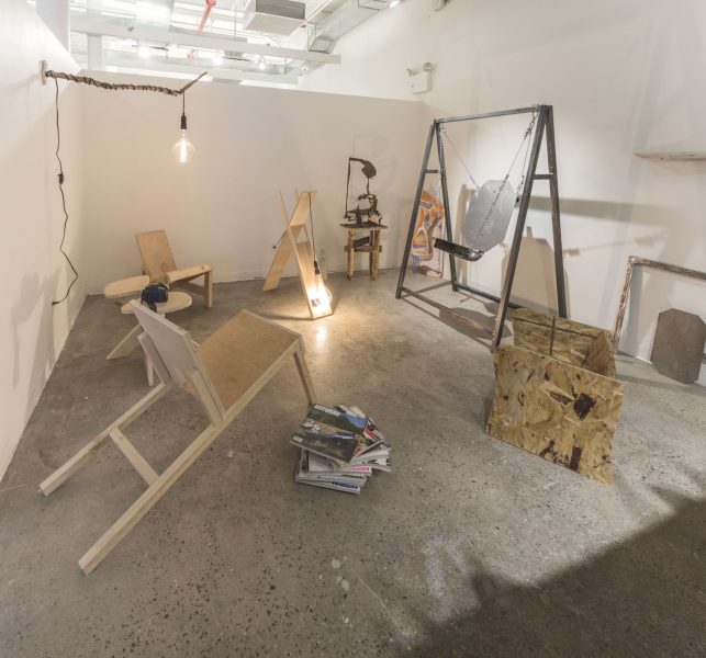 Installation shot of the artist Logan Swedick's studio, with a large plywood chair, a steel swing, and two hanging Edison bulb lamps.