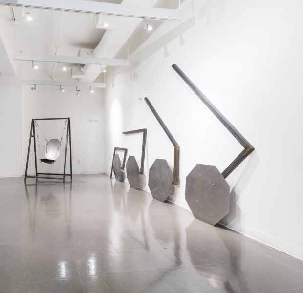 Installation shot of a swing in the background made of metal and four steel sculptures leaning against a wall, looking to be consecutively unfolding.