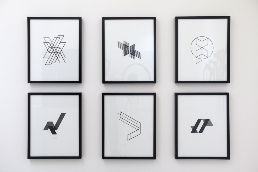 Six framed drawings hang on a white wall. The drawings are each framed with a solid black frame. The drawings each feature different geometrical shapes drawn in black on a white background.