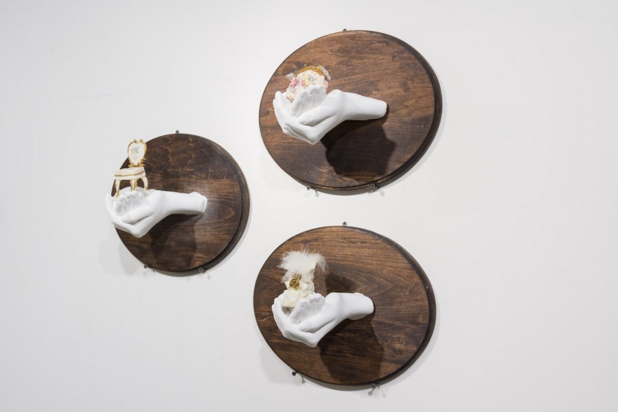 Three cast hands sculptures installed on rounded wood support hung on the wall white the hand sculptures are holding different objects