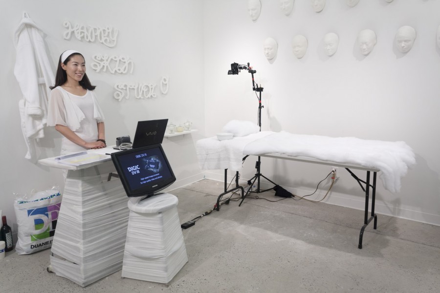 A student standing on a reception desk in a massage room, with a massage table, a computer, and face sculptures installed on the wall
