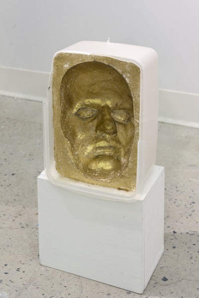 ceramic sculpture representing a face with chin, mouth, nose, and eyes, painted in gold and sitting on a small white stand