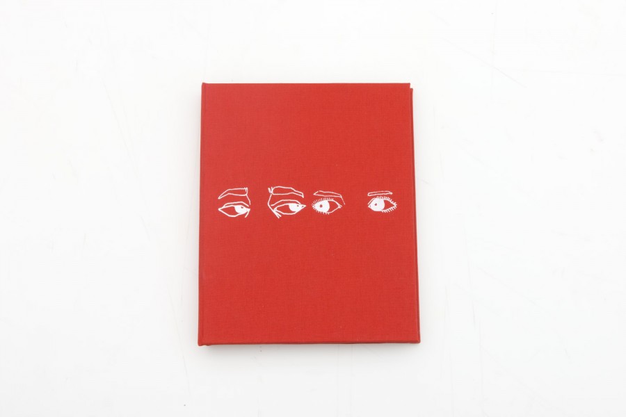 A red booklet on a white table with a drawing of two pairs of eyes and eyebrows made with white ink